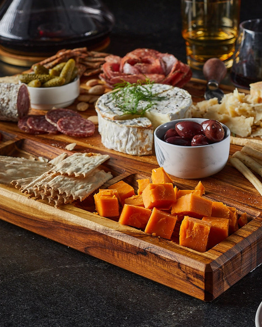 Wooden charcuterie board with various cheeses, meats, and crackers on a dark surface.