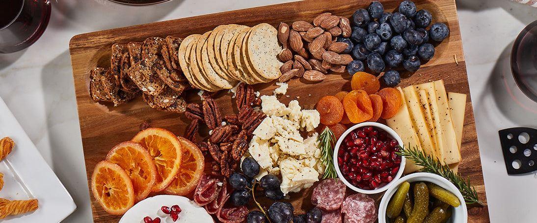Acacia wood charcuterie board with Sonder LA built-in cracker well displaying assorted meats, cheeses, fruits, and crackers on a white surface.