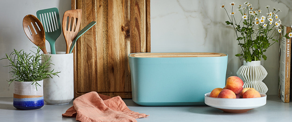 Kitchen scene with a teal Sonder LA bread box, wooden utensils, and a bowl of fruit on a light surface.