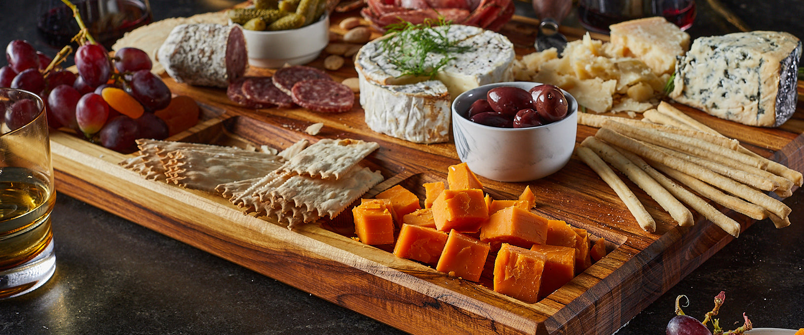 Sonder Los Angeles Bradbury Teak Wood Charcuterie board with built-in compartments serving cheese, grapes, crackers, olives, and sausage. 