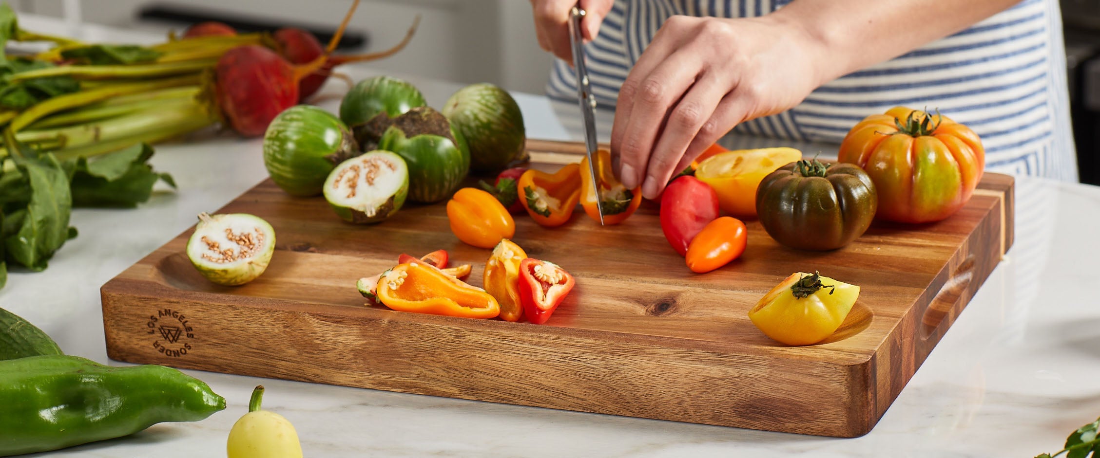 Person cutting vegetables on an acacia wooden cutting board with a variety of peppers and other ingredients.