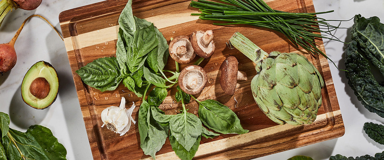 Wooden acacia cutting board with fresh vegetables including artichoke, mushrooms, and herbs on a light background.