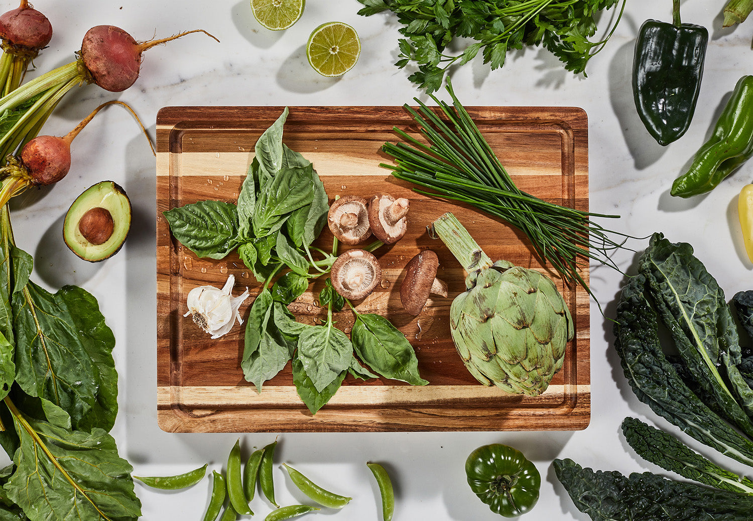 a cutting board with garlic, basil, mushrooms, chives and an artichoke. surrounding the board are limes, beets, avocados, peppers and kale