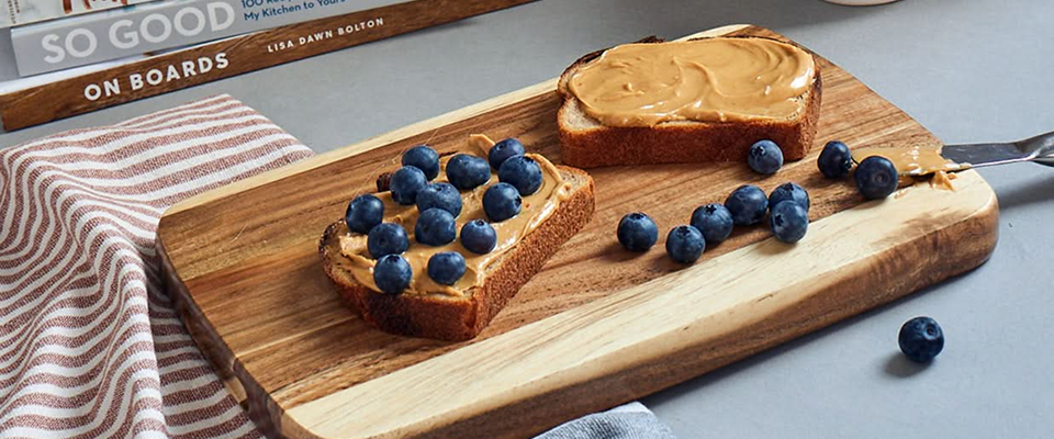 Two slices of toast with peanut butter and blueberries on a Sonder LA wooden cutting board.