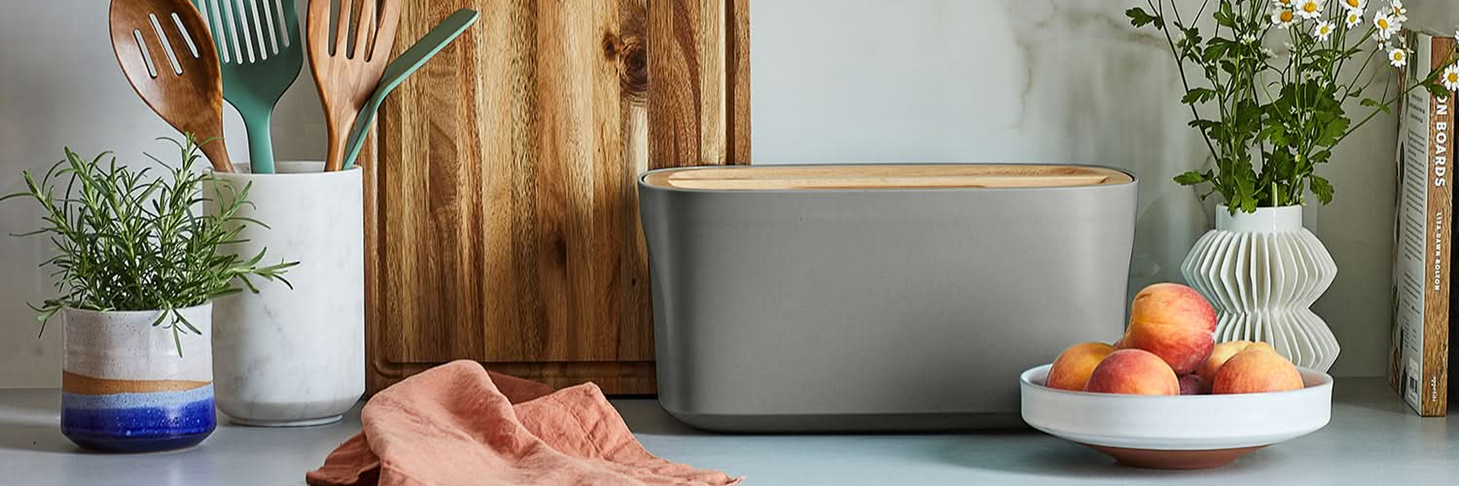 Gray Sonder LA bread box on a kitchen counter with a cutting board, potted plants, and a bowl of fruit.